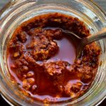 a spoonful of chili oil sits in a glass jar of salsa